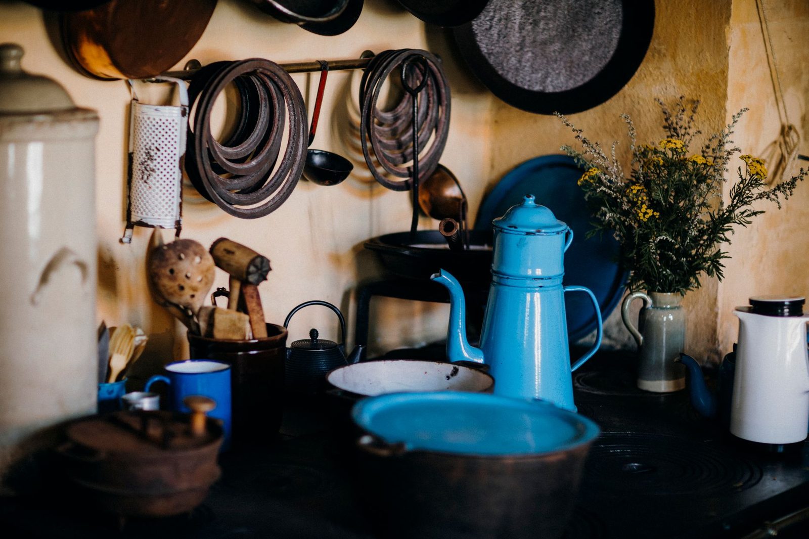 A cozy rustic kitchen features a vintage blue kettle with hanging pans and wildflowers, exuding warmth and charm.